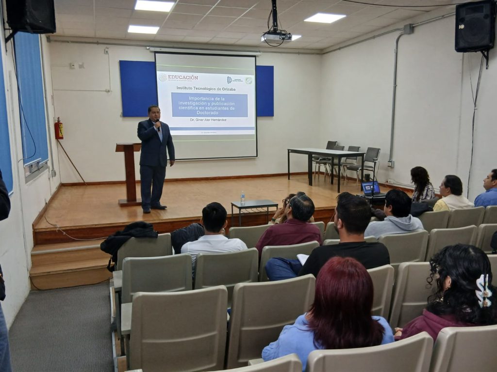 El Dr. Giner Alor Hernández de pie en el escenario, impartiendo la ponencia “Importancia de la Investigación y Publicación Científica en Estudiantes de Doctorado” con una diapositiva proyectada detrás de él.
